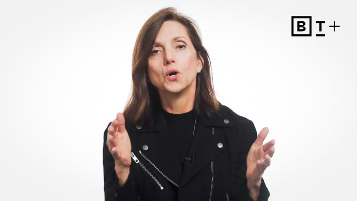 A woman with brown hair in a black jacket speaks with hands raised, against a plain white background with a "BT+" logo in the top right corner.