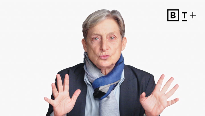 A person with short gray hair, wearing a suit jacket and two-tone scarf, holds both hands up in front of a plain white background with a “B T+” logo in the top right corner.