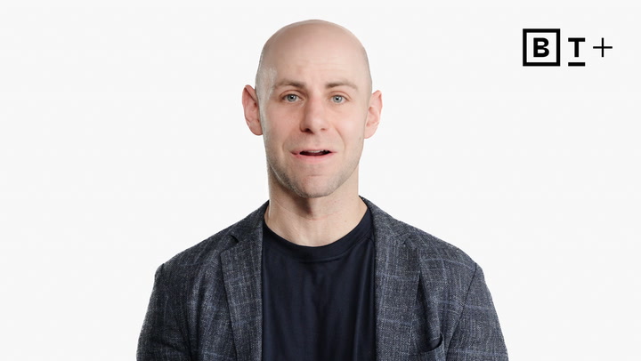 A bald man in a plaid blazer and black shirt stands against a plain white background with a "BT+" logo in the upper right corner.