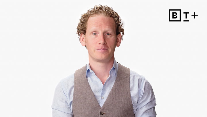 A man with curly hair wearing a light blue shirt and tan vest stands against a white background with a "B T+" logo in the top right corner.