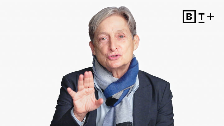 An older person with short gray hair, wearing a suit and a blue scarf, gestures with one hand while speaking. A "B T +" logo is visible in the top right corner.