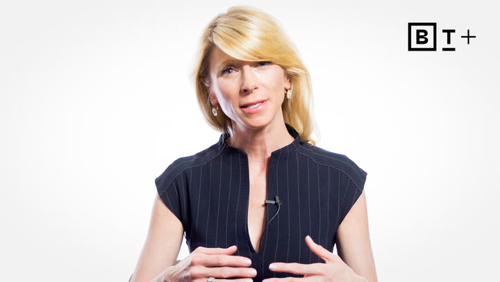 A woman with blonde hair speaks while gesturing with her hands, wearing a dark pinstriped blouse, with a microphone clipped to her shirt and a logo in the top right corner.