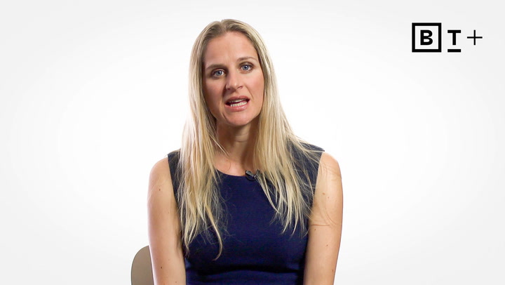 A woman with long blonde hair wearing a navy sleeveless top sits and speaks in front of a plain white background with a "BT+" logo in the top right corner.