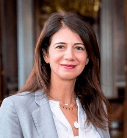 A woman with long brown hair, wearing a light gray blazer and white blouse, stands indoors and smiles at the camera.