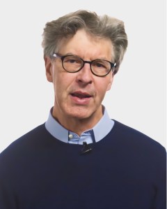 An older man with gray hair and glasses wearing a navy sweater over a light blue collared shirt, speaking in front of a plain white background.