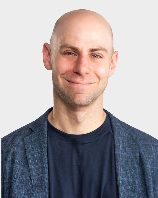 A bald man in a blue blazer and navy shirt smiles at the camera against a plain light background.