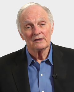 An older man with gray hair wearing a black suit jacket and blue shirt faces the camera with a neutral expression.