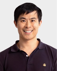 A man with short black hair smiles at the camera, wearing a dark polo shirt with a small embroidered logo on the chest, set against a plain light background.