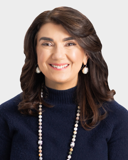 Woman with shoulder-length brown hair, wearing a navy blue sweater, pearl earrings, and a beaded necklace, smiling at the camera against a plain light background.