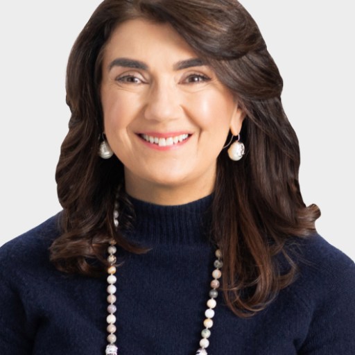 Woman with shoulder-length brown hair, wearing a navy blue sweater, pearl earrings, and a beaded necklace, smiling at the camera against a plain light background.