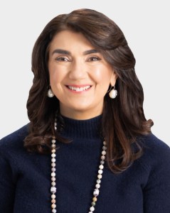 Woman with shoulder-length brown hair, wearing a navy blue sweater, pearl earrings, and a beaded necklace, smiling at the camera against a plain light background.