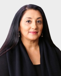 A woman with long dark hair and a neutral expression wears a black top and earrings, posing against a plain light background.