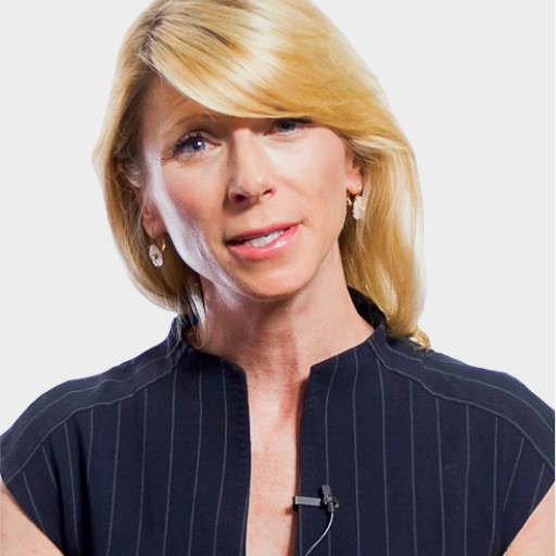 A woman with blonde hair and earrings, wearing a dark, pinstriped top with a microphone clipped to it, speaking against a plain background.