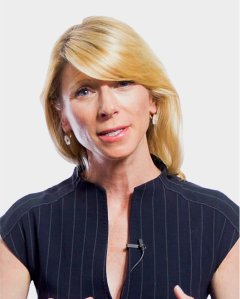 A woman with blonde hair and earrings, wearing a dark, pinstriped top with a microphone clipped to it, speaking against a plain background.