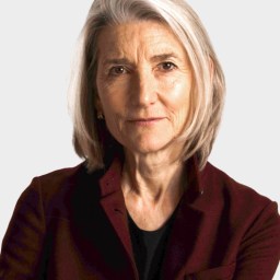 An older woman with shoulder-length gray hair, wearing a dark burgundy jacket over a black top, looks directly at the camera with a neutral expression against a plain background.