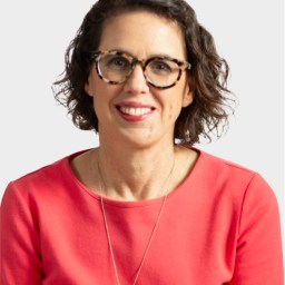 A woman with short, curly brown hair and glasses is wearing a coral top and a necklace, smiling at the camera against a plain light background.