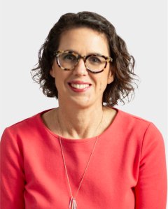 A woman with short, curly brown hair and glasses is wearing a coral top and a necklace, smiling at the camera against a plain light background.