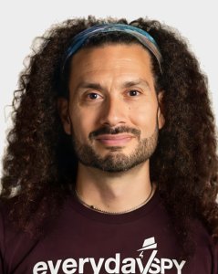 A person with long curly hair, a beard, and a headband, wearing a dark shirt that reads "everyday spy" on a plain background.