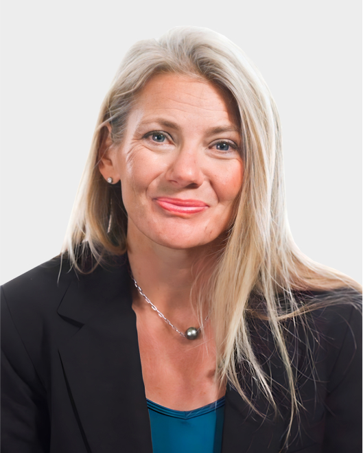 A woman with long blonde hair, wearing a black blazer, blue top, and necklace, poses against a plain light background.