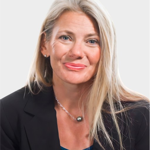 A woman with long blonde hair, wearing a black blazer, blue top, and necklace, poses against a plain light background.