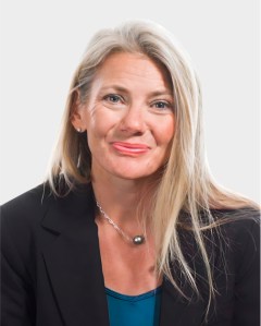 A woman with long blonde hair, wearing a black blazer, blue top, and necklace, poses against a plain light background.