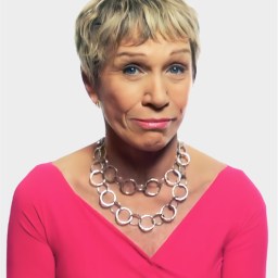 A woman with short blonde hair wears a bright pink top and a chunky silver necklace, looking at the camera with a neutral expression against a plain background.