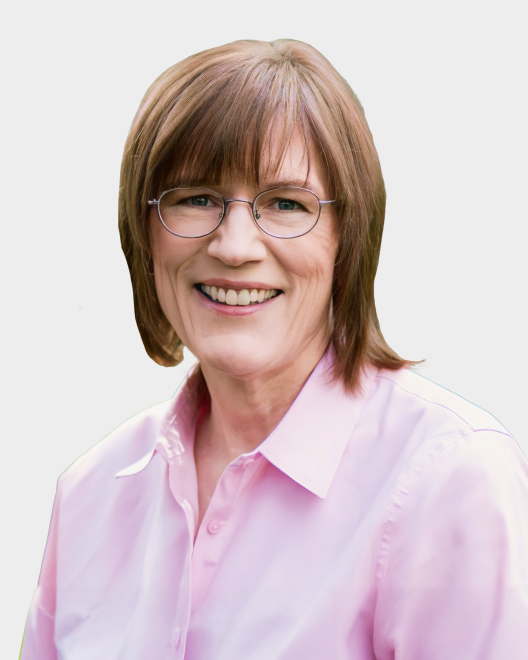 A smiling person with short, light brown hair and glasses, wearing a light pink button-up shirt against a plain background.