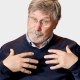 Older man with glasses and gray hair, wearing a dark sweater and collared shirt, gestures toward himself with both hands against a plain light background.