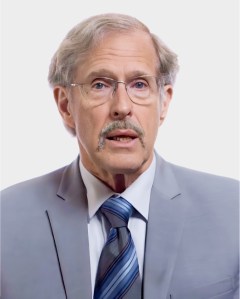 An older man with gray hair, glasses, and a mustache is wearing a light gray suit, white shirt, and striped tie, facing the camera against a plain light background.