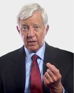 Older man with white hair in a suit and red tie gestures with his right hand while looking forward against a plain background.