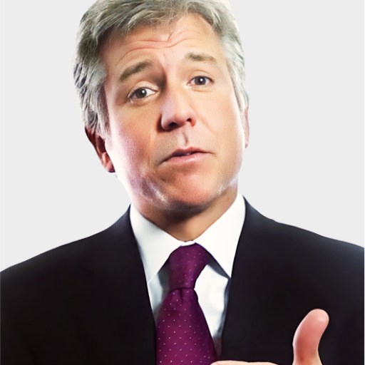 A man in a suit and polka-dot tie gestures with his right hand while speaking, against a plain light background.