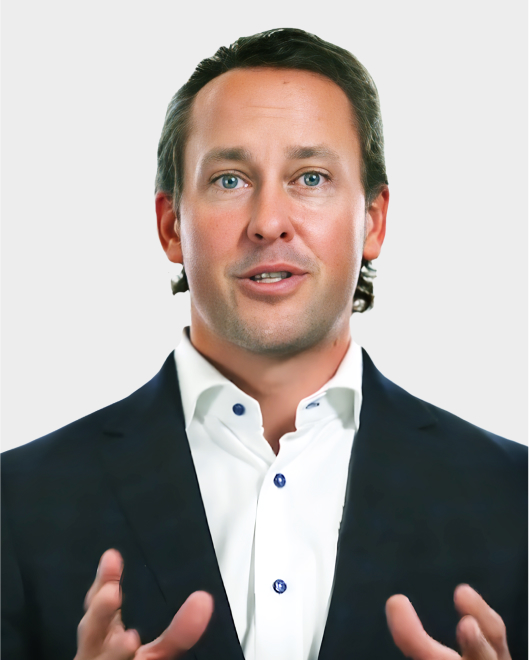 A man in a dark suit and white button-up shirt gestures with his hands while speaking, set against a plain light background.
