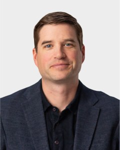 A man with short brown hair wearing a dark blazer and black shirt, facing forward against a plain light background.
