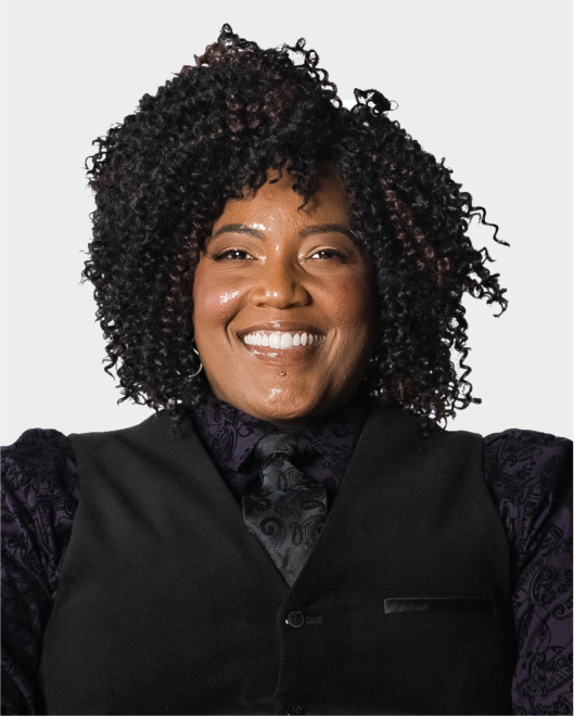 A person with curly hair, wearing a patterned dark shirt, vest, and tie, smiling in front of a plain light background.