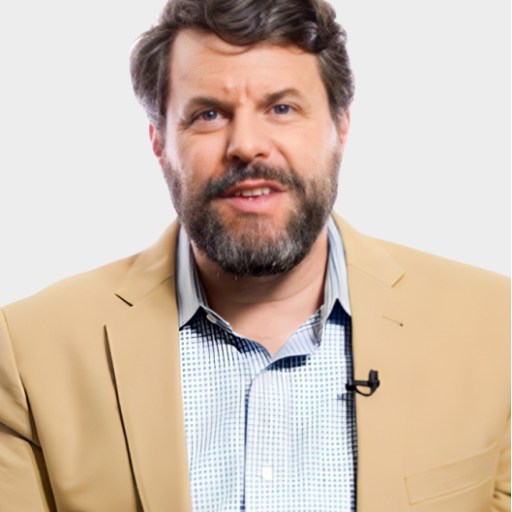 A man with a brown beard and hair, wearing a tan blazer over a checked shirt, speaks while facing the camera against a plain light background.