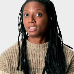 A person with long braided hair wearing a beige knit sweater and a pendant necklace, looking slightly to the side against a plain background.