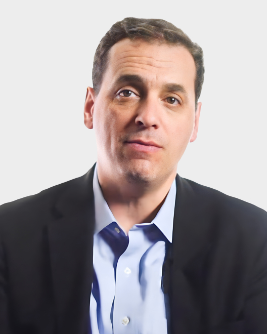 A man with short dark hair wearing a blue shirt and dark blazer poses in front of a plain light background.