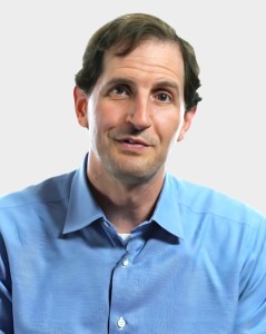 A man with short brown hair wearing a light blue button-up shirt poses against a plain light background.