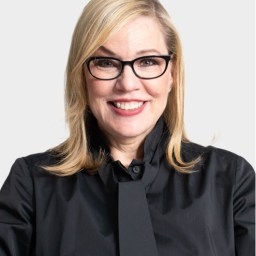 A woman with blonde hair and black-framed glasses smiles at the camera, wearing a black button-up shirt and tie against a plain light background.