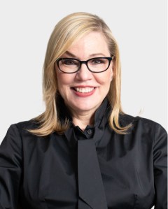 A woman with blonde hair and black-framed glasses smiles at the camera, wearing a black button-up shirt and tie against a plain light background.