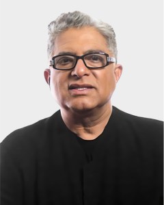 A middle-aged person with short gray hair and glasses, wearing a black shirt, is facing forward against a plain light background.