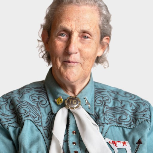 An older woman with gray hair wears a teal embroidered shirt, a white scarf with a bolo tie, and looks directly at the camera against a plain background.