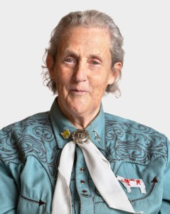 An older woman with gray hair wears a teal embroidered shirt, a white scarf with a bolo tie, and looks directly at the camera against a plain background.