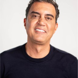 A man with short curly hair wearing a black crew-neck shirt is smiling at the camera against a plain, light background.