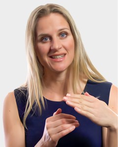 A woman with long blonde hair and a navy sleeveless top gestures with her hands while smiling at the camera against a plain light background.