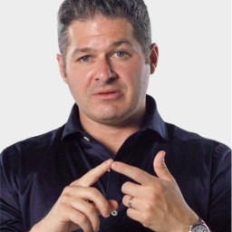 A man with short dark hair in a dark shirt gestures with his hands, pointing with his index fingers, against a plain light background.