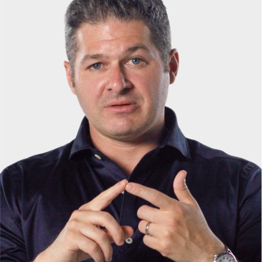 A man with short dark hair in a dark shirt gestures with his hands, pointing with his index fingers, against a plain light background.