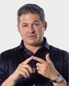 A man with short dark hair in a dark shirt gestures with his hands, pointing with his index fingers, against a plain light background.