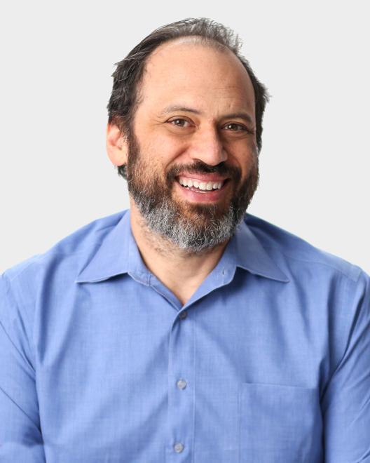 A man with dark hair and a beard, wearing a light blue collared shirt, smiles at the camera against a plain light background.