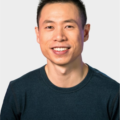A man with short black hair, wearing a dark blue crew neck shirt, smiling at the camera against a plain light background.